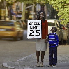 A woman holds a 25MPH speed limit sign with one hand and with the other she holds her child's hand, who's looking up at her.